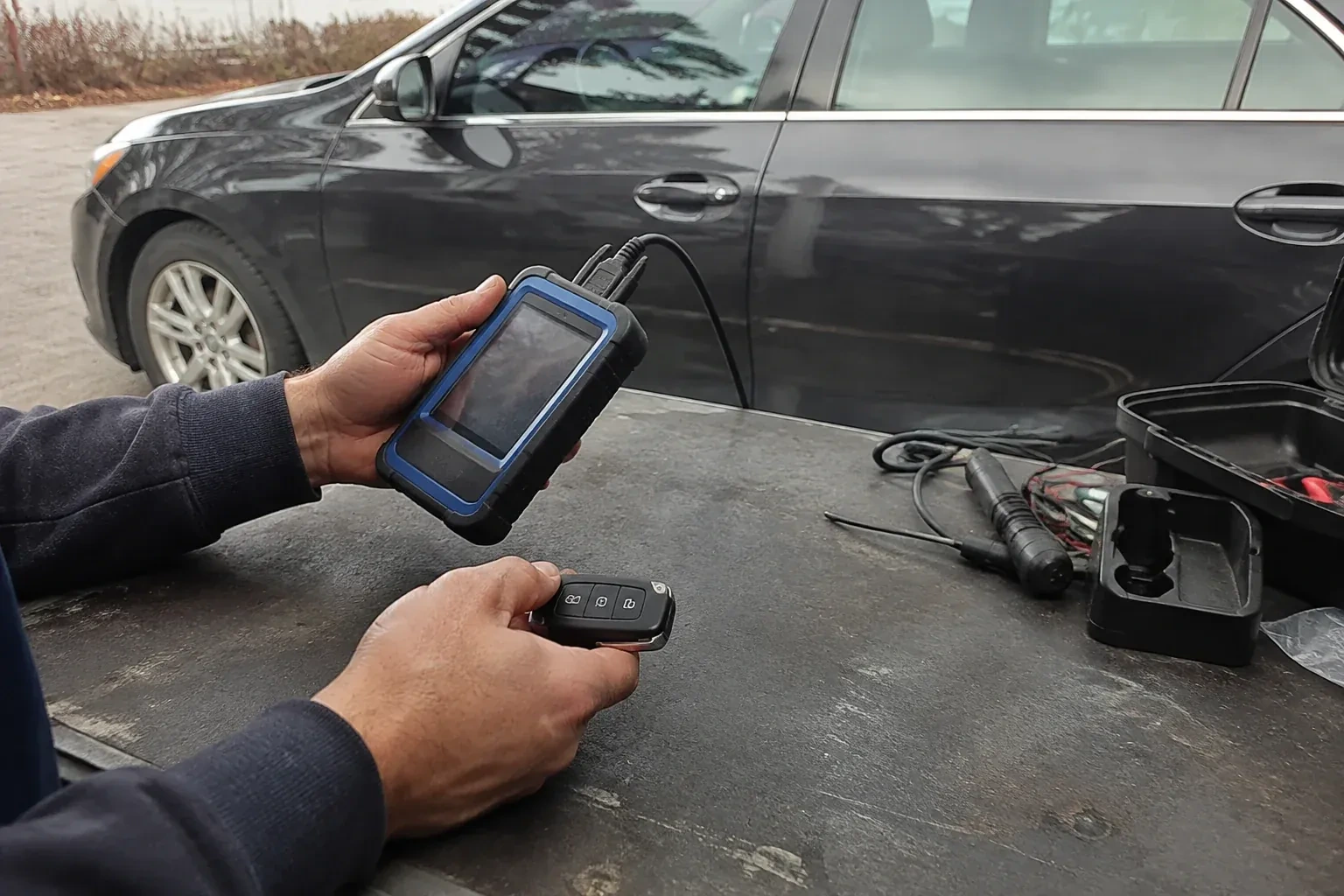 Hands holding a blue-edged electronic device and car-key fob at a worktable beside a parked car. — Transponder Key Programming, Hamilton