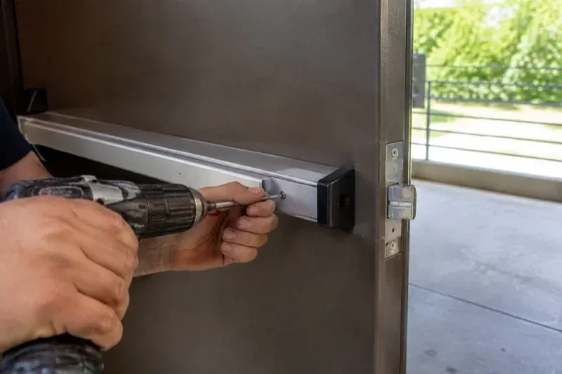Hands using a power driver at the end of a horizontal push bar on an open metal door. — Panic & Exit Hardware, Hamilton